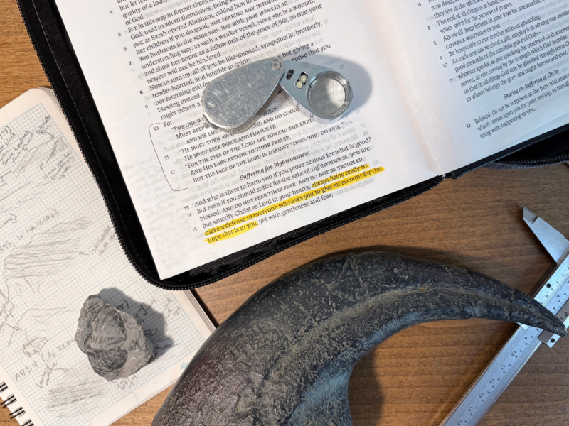 A Bible on a table opened to 1 Peter 3:15 surrounded by a geology journal, fossils, a hand lens, and calipers.