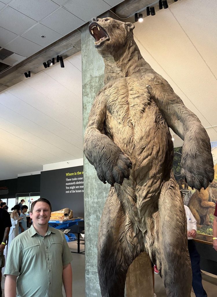 A life-sized reconstruction of the extinct short-faced bear standing on its hind legs. A man standing next to it only reaches up to the bear's waist.